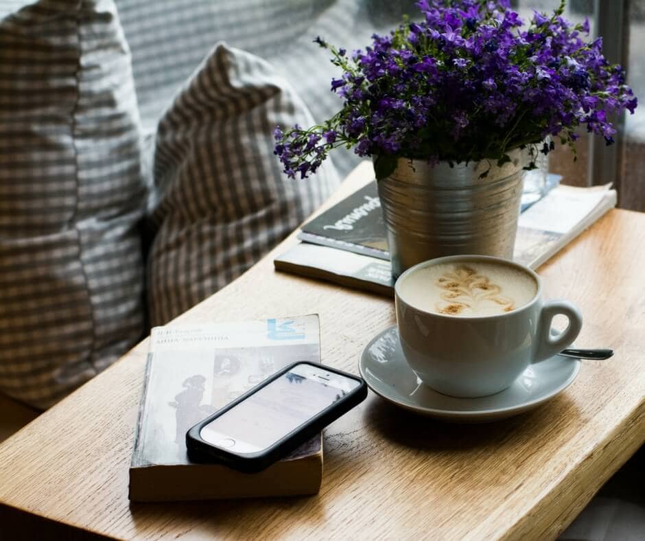 A cozy setting with a cup of cappuccino, a book, a smartphone, and a pot of purple flowers on a wooden table near checkered pillows, perfect for planning your next post using a social media planner | plannthat.com