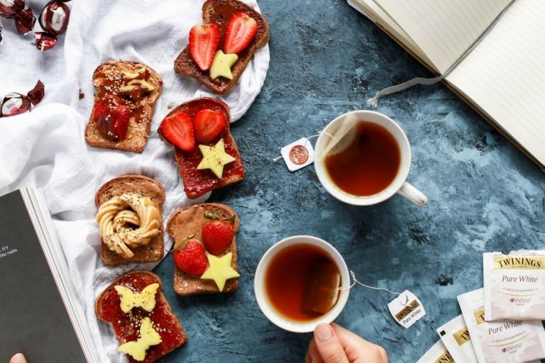 A cozy breakfast scene with assorted toast topped with strawberries and butter, two cups of tea, a notebook, chocolates, and Twinings tea bags on a blue textured surface, perfect for planning your day with a social media scheduler | plannthat.com