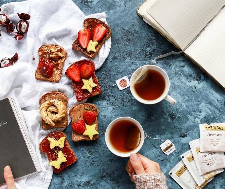 A cozy breakfast scene with assorted toast topped with strawberries and butter, two cups of tea, a notebook, chocolates, and Twinings tea bags on a blue textured surface, perfect for planning your day with a social media scheduler | plannthat.com