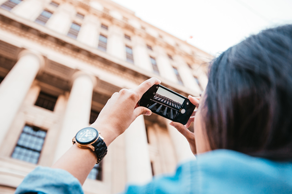 A person with long hair and a wristwatch takes a photo of a grand building with tall columns using a smartphone, focusing on the building's architectural details from behind | Social media planner | plannthat.com