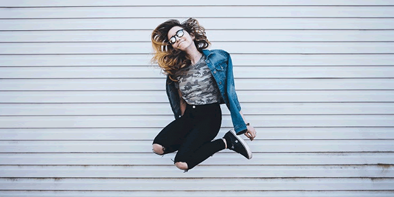 A woman with long hair, wearing glasses, a denim jacket, and black pants, jumps joyfully in front of a white, horizontally-striped wall, capturing a moment of carefree excitement with an Instagram scheduler | plannthat.com