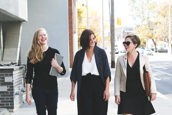 Three women walking and smiling on a sunny sidewalk, dressed in business casual attire, carrying bags and a laptop, perfect for a social media planner | plannthat.com