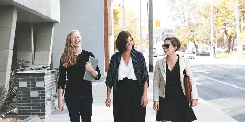 Three women walking and smiling on a sunny sidewalk, dressed in business casual attire, carrying bags and a laptop, perfect for a social media planner | plannthat.com