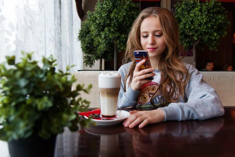 A young woman with long, wavy hair sits at a café table, looking at her smartphone, possibly using an Instagram scheduler. A latte with foam and a small plant are on the table, with greenery in the background. | plannthat.com