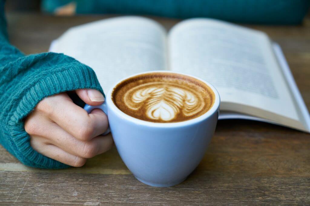 A person in a teal sweater holding a cup of latte with intricate latte art, sitting at a wooden table with an open book in the background, perfect for a social media planner | plannthat.com