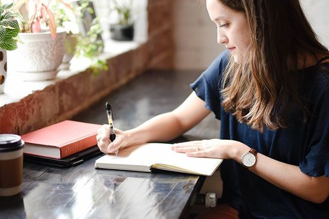 A woman writing in a notebook at a wooden table near a window with plants, with a coffee cup and a book also on the table, using her social media planner | plannthat.com