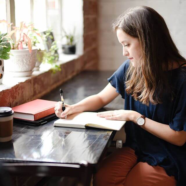 A woman writing in a notebook at a wooden table near a window with plants, with a coffee cup and a book also on the table, using her social media planner | plannthat.com