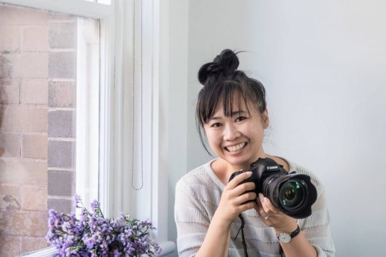 A smiling woman with a topknot hairstyle holds a Canon camera while sitting by a window with purple flowers, wearing a light-colored sweater against a white wall, creating a bright and cheerful atmosphere | Social media planner | plannthat.com