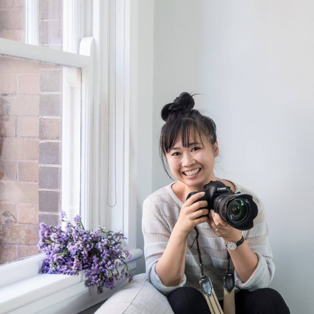 A smiling woman with a topknot hairstyle holds a Canon camera while sitting by a window with purple flowers, wearing a light-colored sweater against a white wall, creating a bright and cheerful atmosphere | Social media planner | plannthat.com