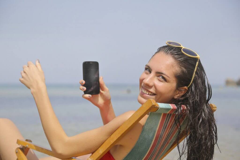 A woman with wet hair, wearing sunglasses, smiles while holding a smartphone. She is lounging on a striped beach chair by the sea, enjoying a sunny day. The background features a clear blue sky and calm ocean waters, perfect for using a social media scheduler | plannthat.com