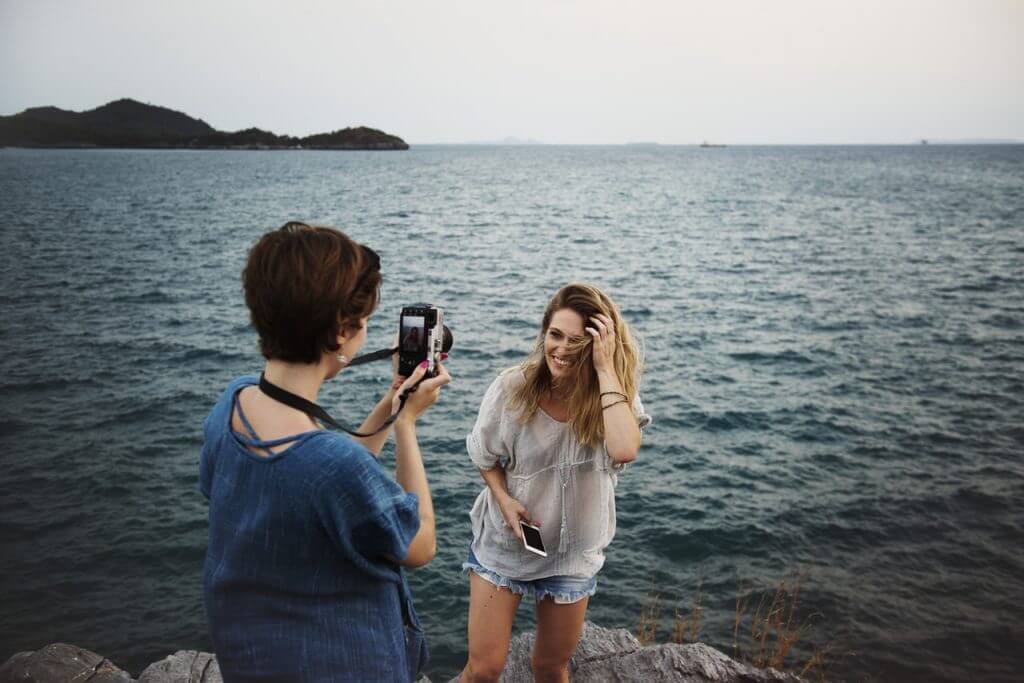 Two people at a seaside cliff, one taking a photo of the other with a camera, against a backdrop of the ocean and distant islands, capturing moments for their Instagram scheduler | plannthat.com