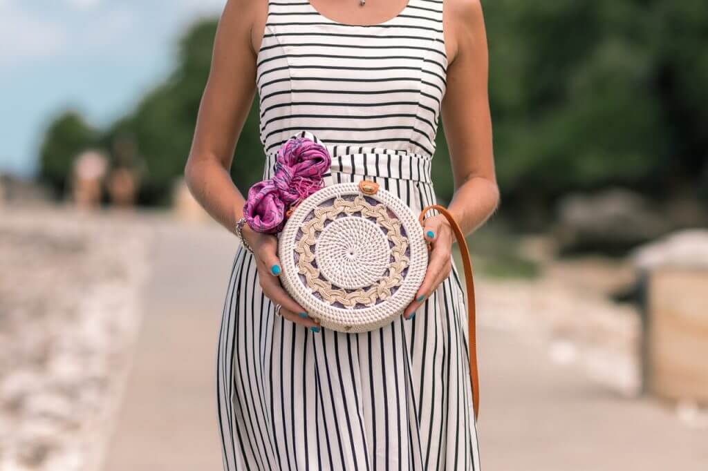 A woman in a striped dress holds a round, woven handbag with a pink scarf tied to it, standing on a pathway with greenery and a cloudy sky in the background, showcasing a stylish look for your Instagram scheduler | plannthat.com