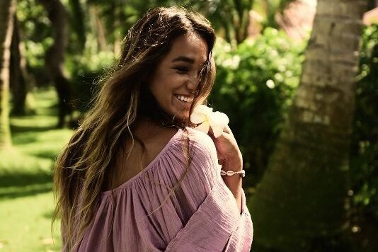 A woman with long hair, wearing a pink off-shoulder top, smiles while looking back, surrounded by lush greenery and palm trees in a tropical atmosphere with sunlight filtering through the leaves, casting a warm glow, perfect for a social media planner | plannthat.com