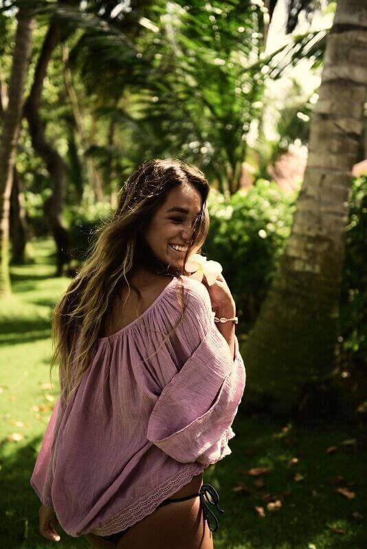 A woman with long hair, wearing a pink off-shoulder top, smiles while looking back, surrounded by lush greenery and palm trees in a tropical atmosphere with sunlight filtering through the leaves, casting a warm glow, perfect for a social media planner | plannthat.com