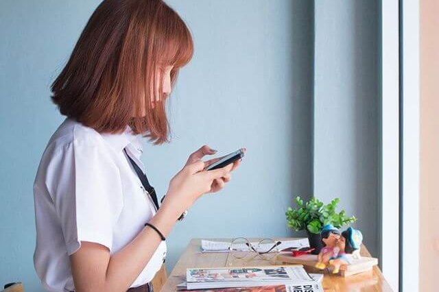A woman in a white shirt looks at her phone while standing at a wooden table with magazines, glasses, and a small plant by a window, possibly using a social media scheduler | plannthat.com