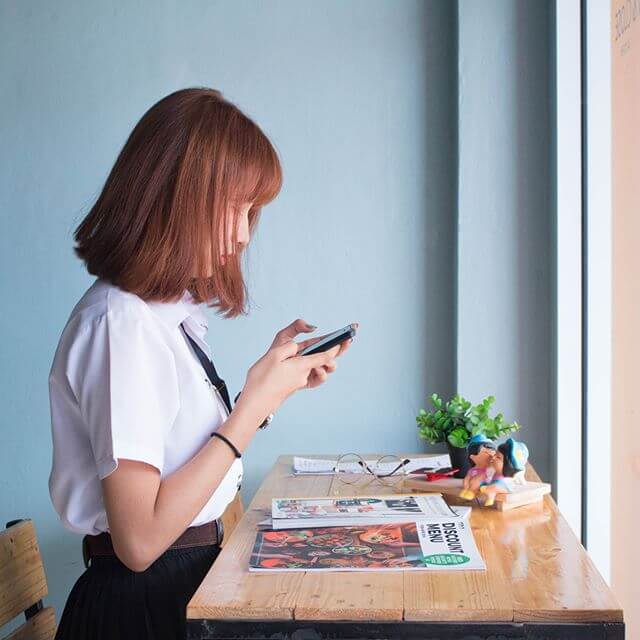 A woman in a white shirt looks at her phone while standing at a wooden table with magazines, glasses, and a small plant by a window, possibly using a social media scheduler | plannthat.com