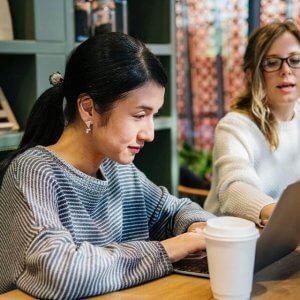 Two women working together at a table with a laptop and coffee cup, one typing while the other points at the screen in a modern, well-lit workspace, using a social media scheduler | plannthat.com