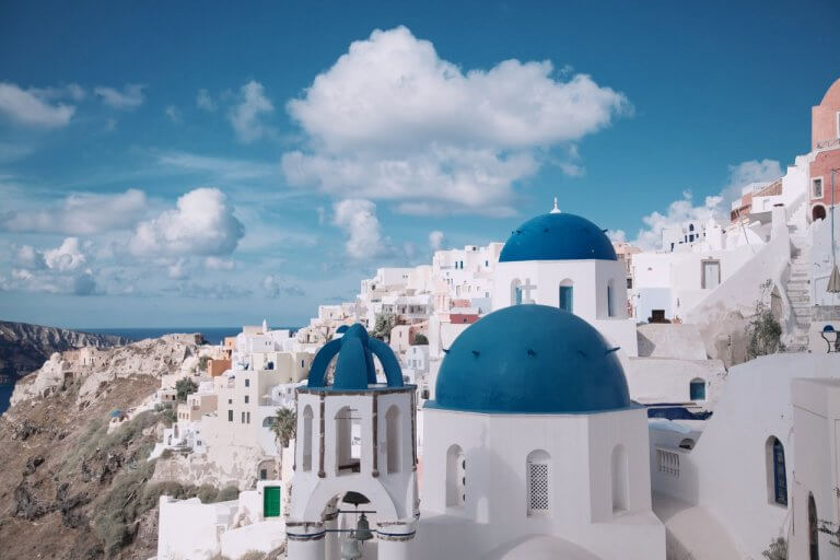 A picturesque view of Santorini, Greece, featuring white-washed buildings with iconic blue domes under a bright blue sky with scattered clouds, with the Aegean Sea in the background, perfect for an Instagram scheduler | plannthat.com