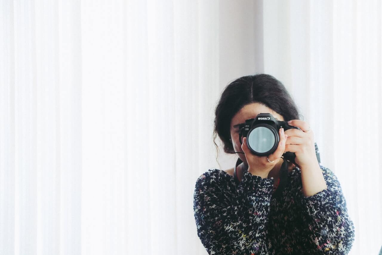 A person with dark hair, wearing a patterned sweater, is holding a camera up to their face, taking a photo in front of a white curtain background, creating a bright and minimalist setting, perfect for a social media planner | plannthat.com