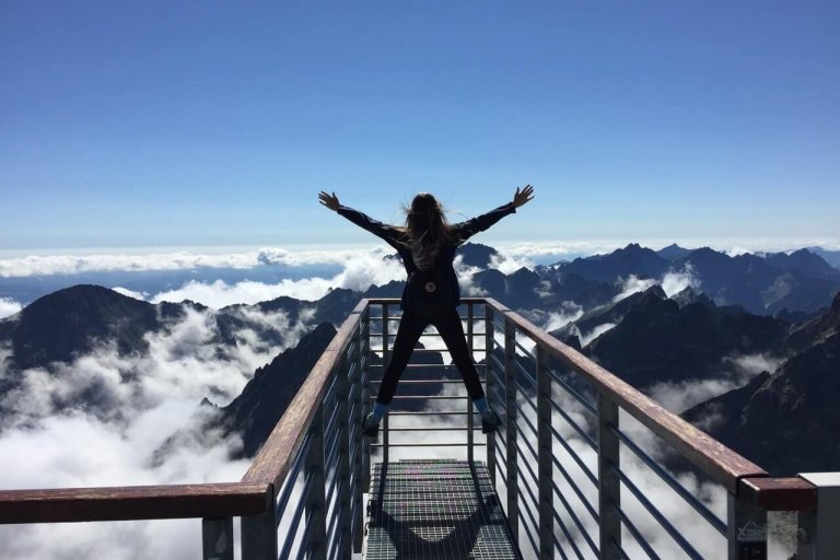Person standing on a mountain viewing platform, arms raised, overlooking a scenic landscape of clouds and distant peaks under a clear blue sky, perfect for planning content with an Instagram scheduler | plannthat.com