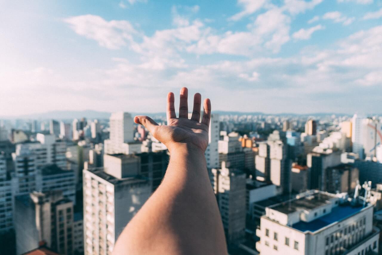 A person's outstretched arm and hand reaching towards a cityscape with numerous buildings under a blue sky with scattered clouds, suggesting aspiration or connection with the urban environment from a high vantage point, highlighting the potential of using a social media planner | plannthat.com