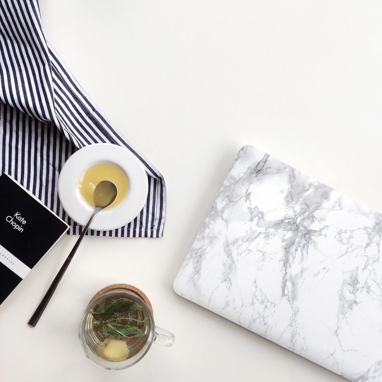 "A marble-patterned laptop, striped cloth, book, honey in a dish with spoon, and herbal tea on a white surface, creating a cozy workspace for using a social media scheduler" | plannthat.com