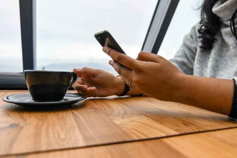 Person using a smartphone while sitting at a wooden table with a black coffee cup, next to a large window, setting up an Instagram business profile | plannthat.com