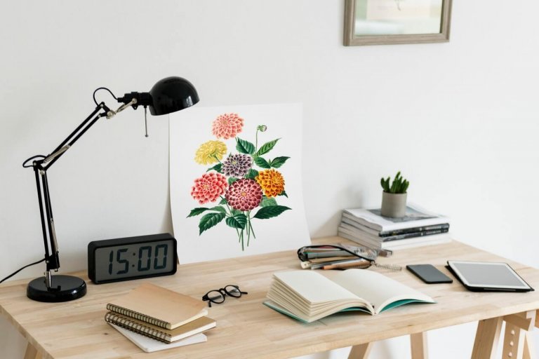 A tidy wooden desk with a lamp, digital clock, notebooks, glasses, tablet, and a floral art print, perfect for a social media planner. A small plant and stack of books are in the background. | plannthat.com