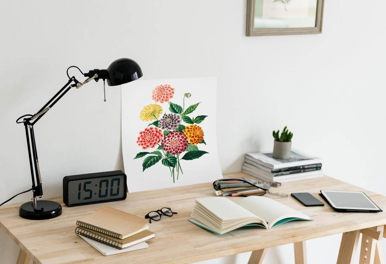 A tidy wooden desk with a lamp, digital clock, notebooks, glasses, tablet, and a floral art print, perfect for a social media planner. A small plant and stack of books are in the background. | plannthat.com