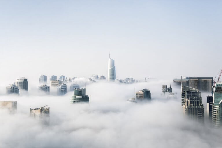 A city skyline with modern skyscrapers emerging through a thick layer of fog under a clear blue sky, with the tallest building centrally located and surrounded by various other high-rise structures, creating a serene and ethereal scene, perfect for a social media planner | plannthat.com