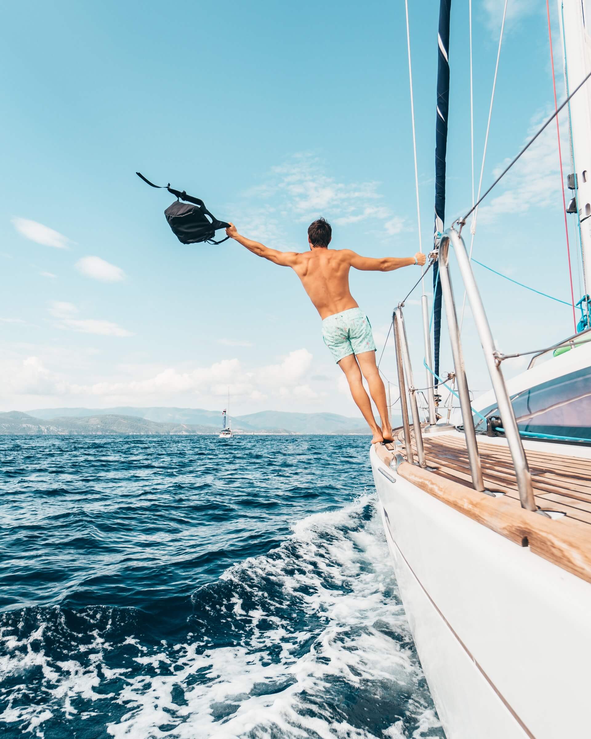 A man in swim trunks stands on the edge of a sailboat, holding a bag and extending his arm outwards over the ocean with a clear sky and another sailboat in the distance, enjoying a moment of freedom and adventure | Social media scheduler | plannthat.com