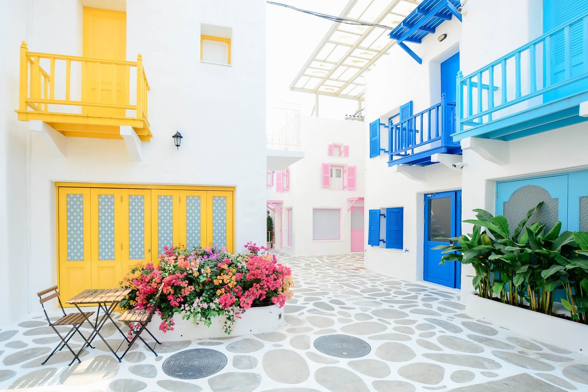 A vibrant courtyard with white buildings featuring yellow, blue, and pink doors and balconies, adorned with colorful flowers and plants, along with a small table and chairs, creating a charming ambiance, perfect for capturing with an Instagram scheduler | plannthat.com