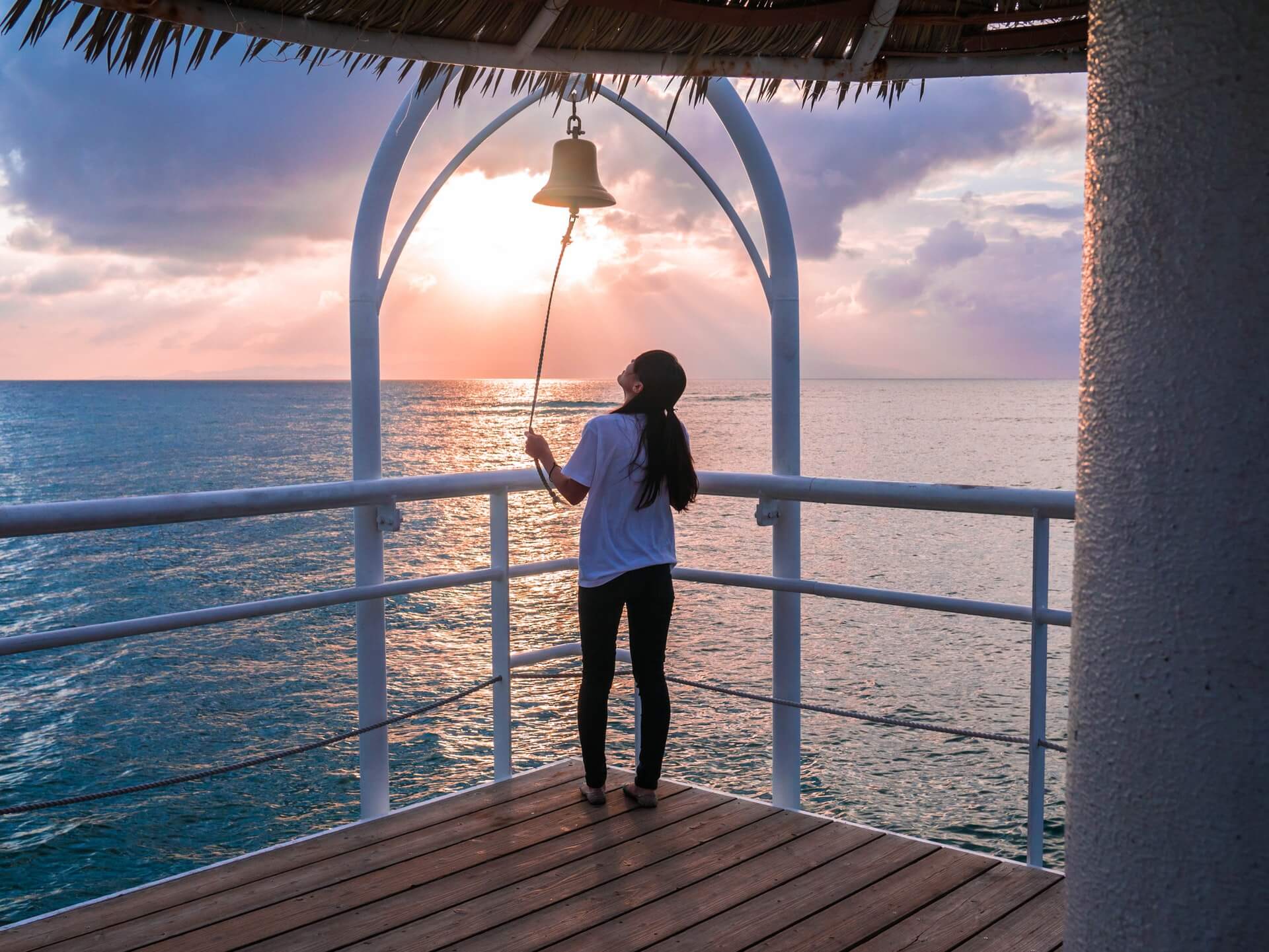 A person stands on a wooden deck by the ocean, ringing a bell at sunset with clouds filling the sky and a warm glow on the water | Enhance your Instagram scheduler experience | plannthat.com