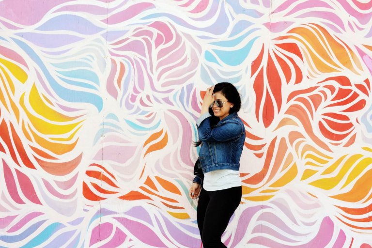 A woman in a denim jacket and sunglasses smiles while standing against a colorful, abstract mural with swirls of red, purple, blue, yellow, and orange, showcasing her vibrant personality | Social media planner | plannthat.com