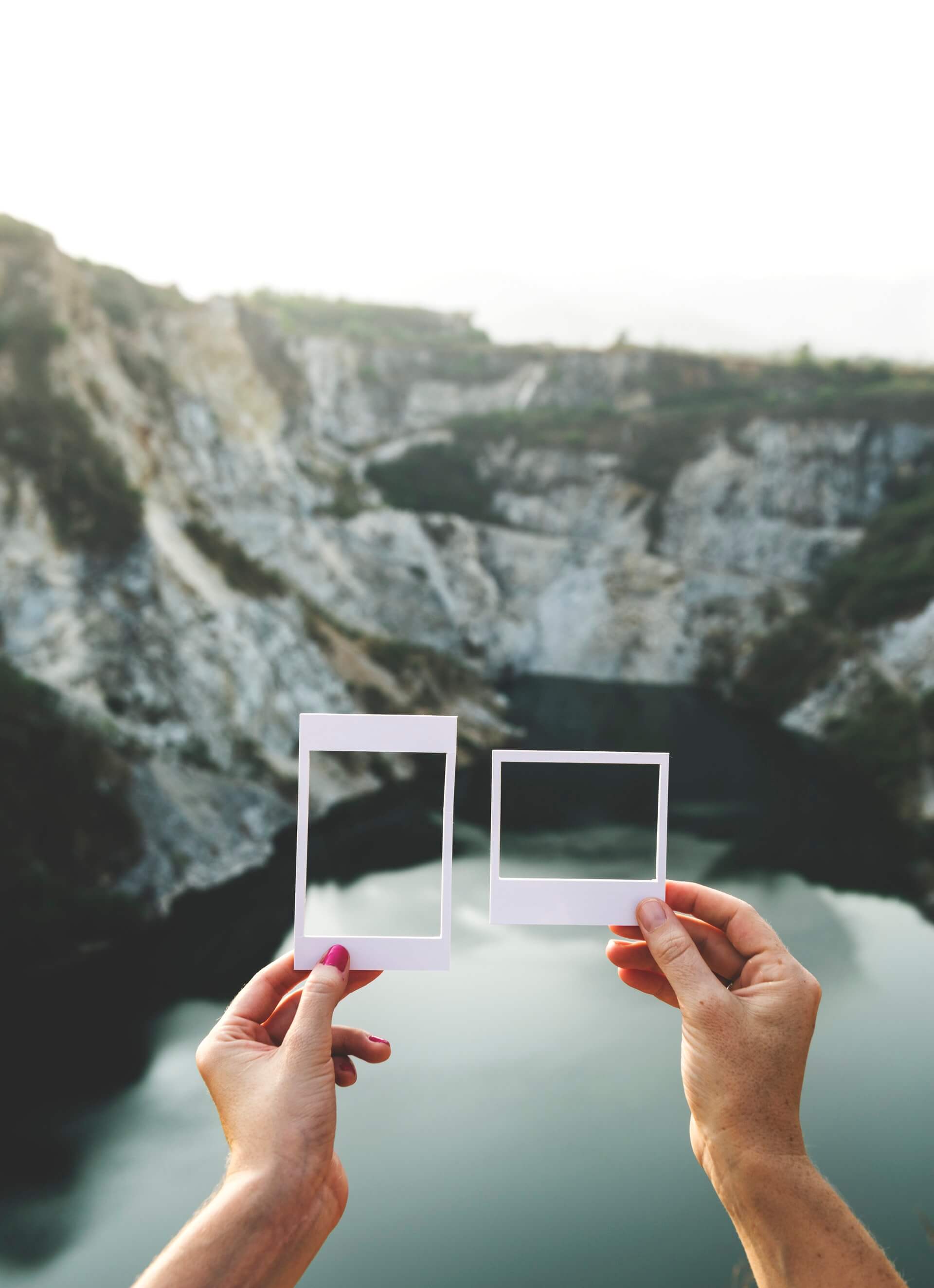 Two hands holding empty Polaroid frames against a scenic backdrop of a rocky cliff and a calm lake, creatively capturing parts of the landscape | Social media scheduler | plannthat.com