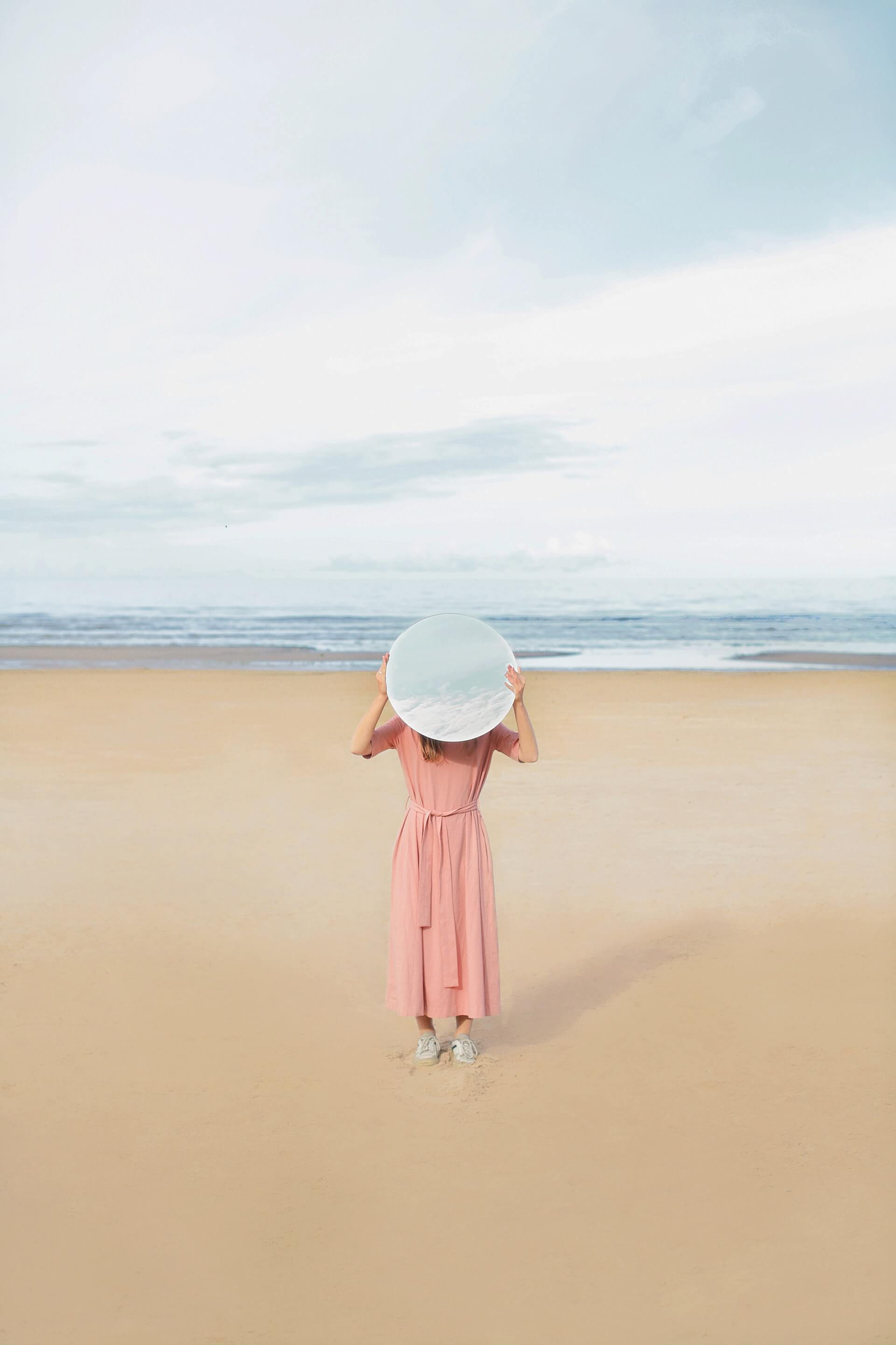 Person in a pink dress holding a round mirror, standing on a sandy beach with the ocean and cloudy sky in the background, showcasing the journey behind the scenes of using a social media scheduler | plannthat.com