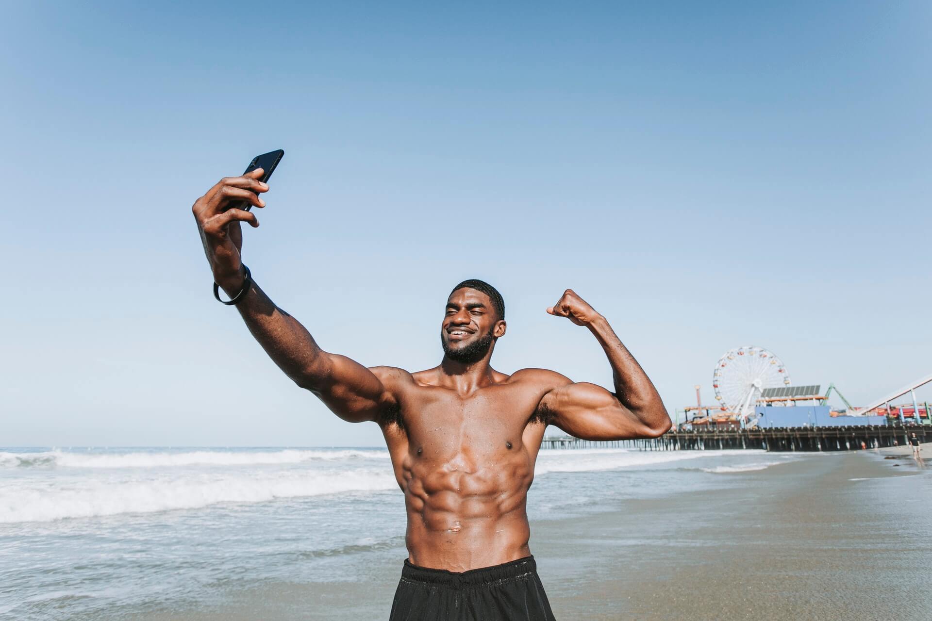 Man flexing muscles and taking a selfie at the beach, with a pier and Ferris wheel in the background on a clear, sunny day, showcasing his fitness journey for his followers using an Instagram scheduler | plannthat.com