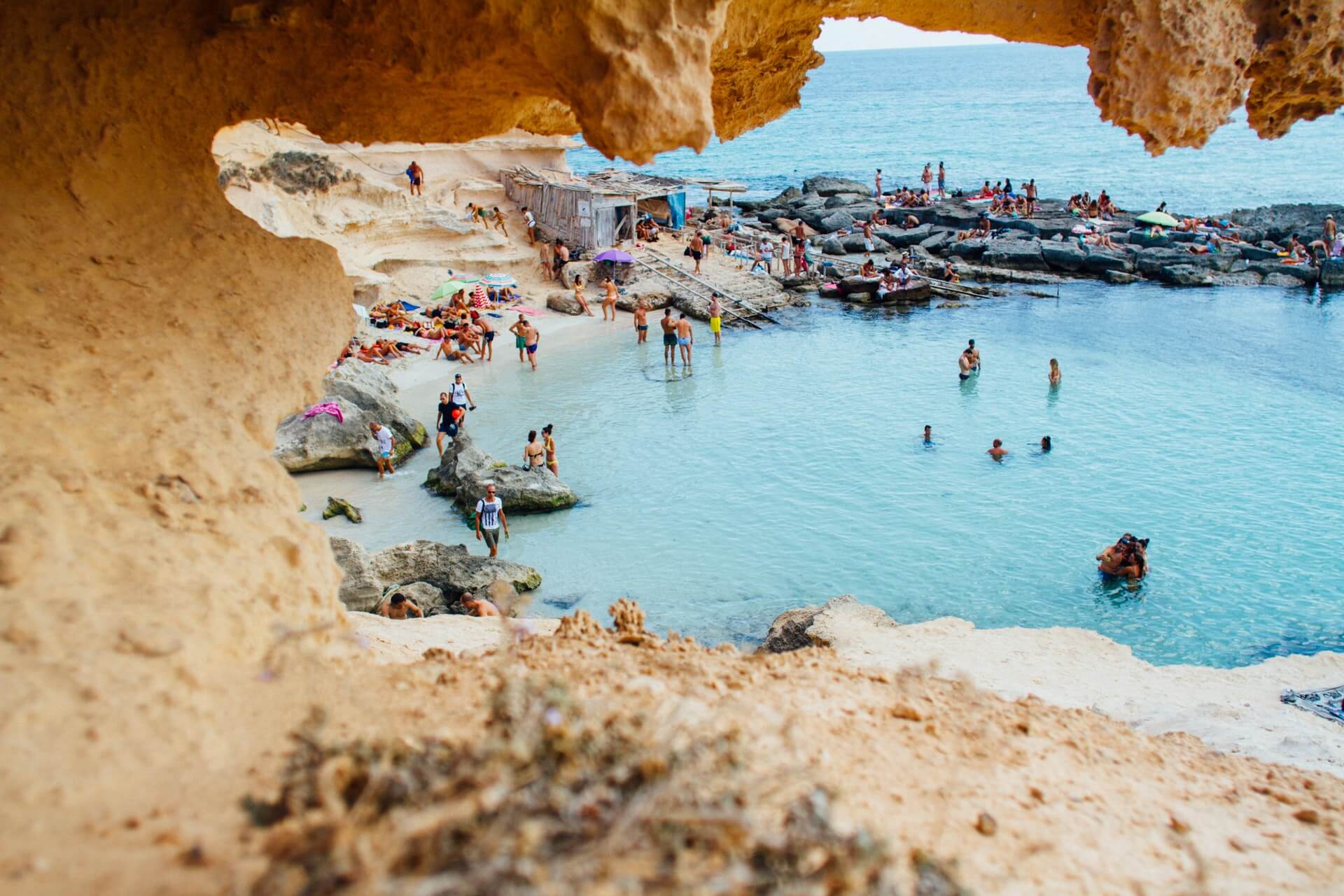 A scenic beach view framed by rocky cliffs, showing people swimming, sunbathing, and relaxing by the clear blue water on a lively, sunny day at a popular coastal destination with natural rock formations and vibrant activity, perfect for capturing moments with an Instagram scheduler | plannthat.com