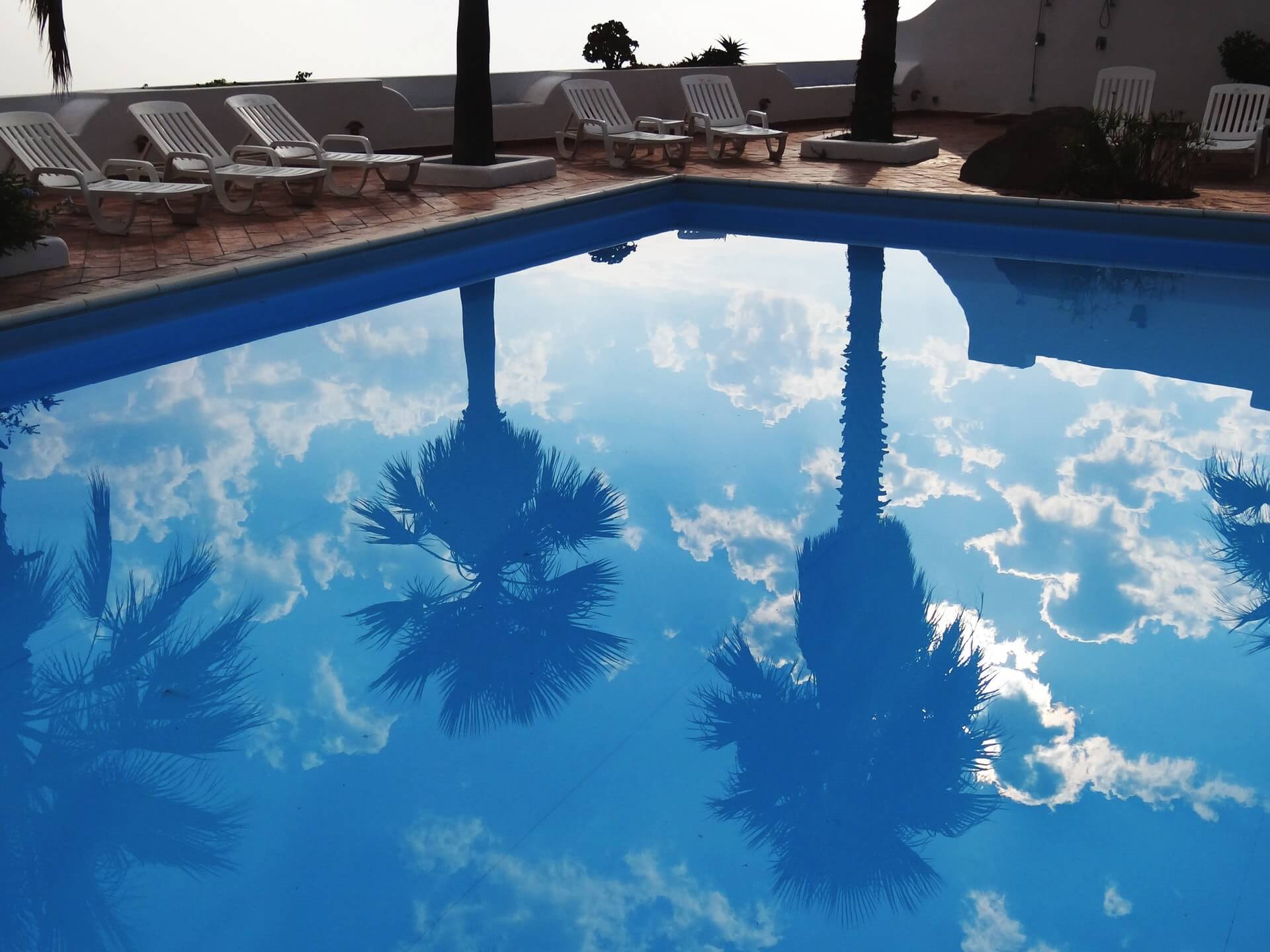 Reflection of palm trees and clouds in a clear blue swimming pool, surrounded by empty lounge chairs on a tiled deck, perfect for planning your next Instagram post with a social media scheduler | plannthat.com
