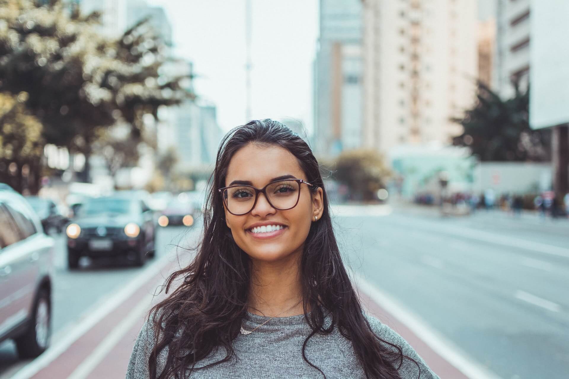 A woman with glasses and long hair smiles while standing on a city street with blurred buildings and cars in the background, showcasing her role as a social media planner | plannthat.com