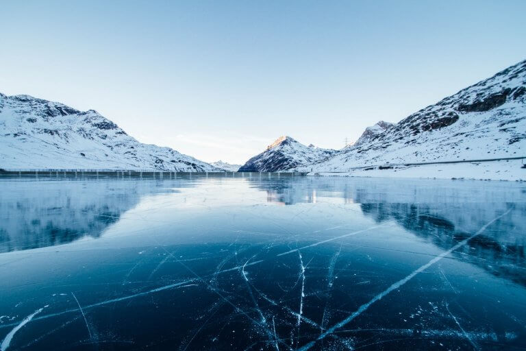 A serene frozen lake surrounded by snow-covered mountains under a clear blue sky, with reflections and ice cracks visible on the lake's surface, perfect for capturing with an Instagram scheduler | plannthat.com