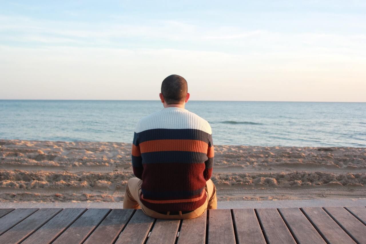 A person in a striped sweater sits on a wooden platform, facing a serene beach and calm ocean, under a clear sky, using a social media planner to organize content | plannthat.com