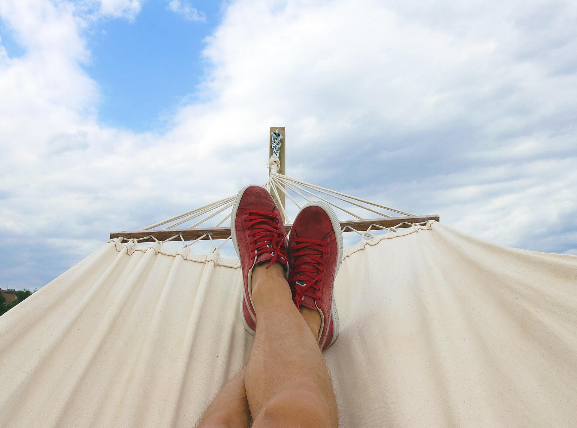 Person relaxing on a hammock with legs crossed, wearing red shoes, against a partly cloudy sky backdrop, stress-less-instagram-marketing | Instagram scheduler | plannthat.com