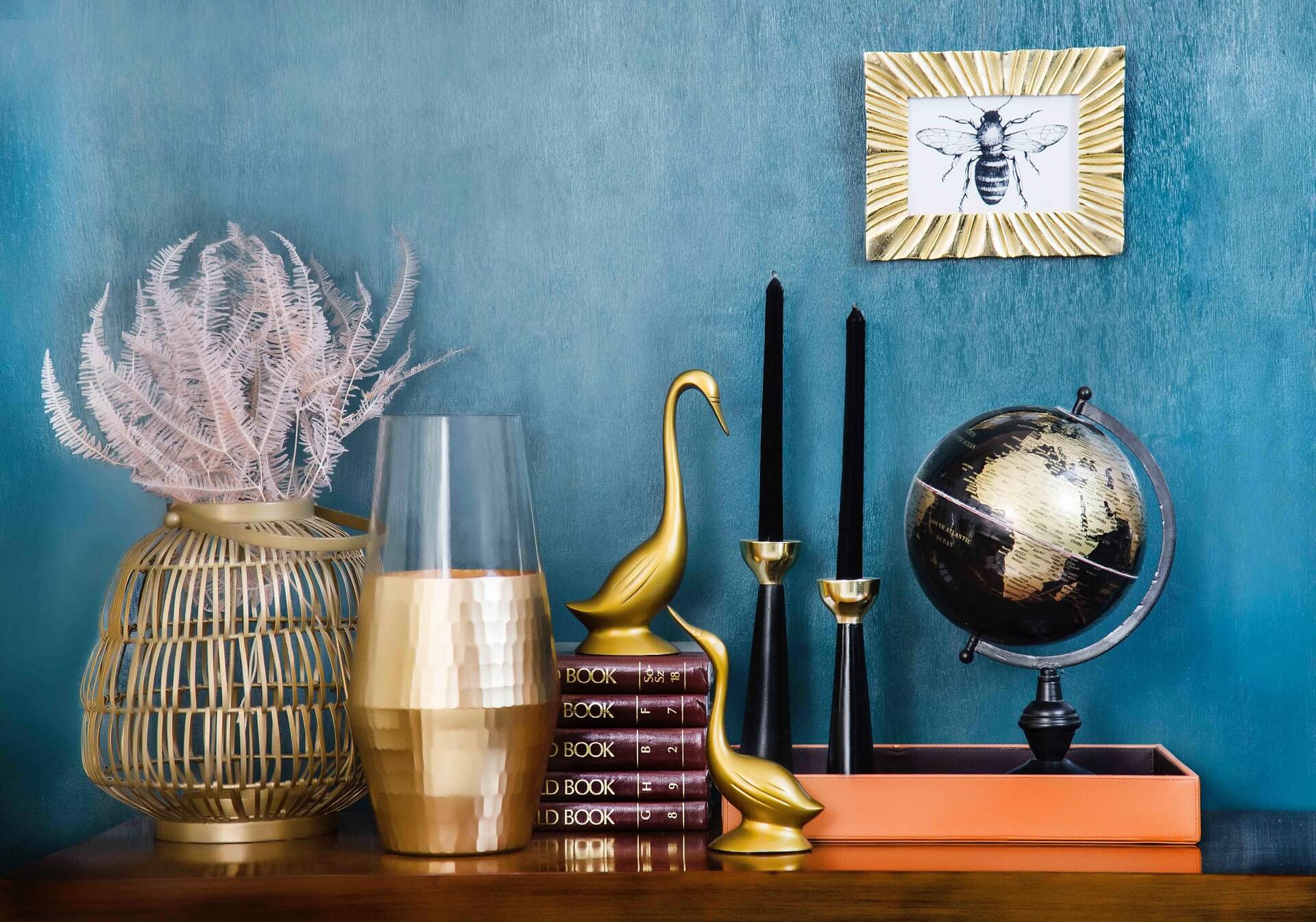 A stylish vignette with gold accents, featuring a globe, black candles, gold bird statues, books, a framed bee print, and a decorative vase with pink foliage against a blue textured wall, perfect for a social media scheduler setup | plannthat.com