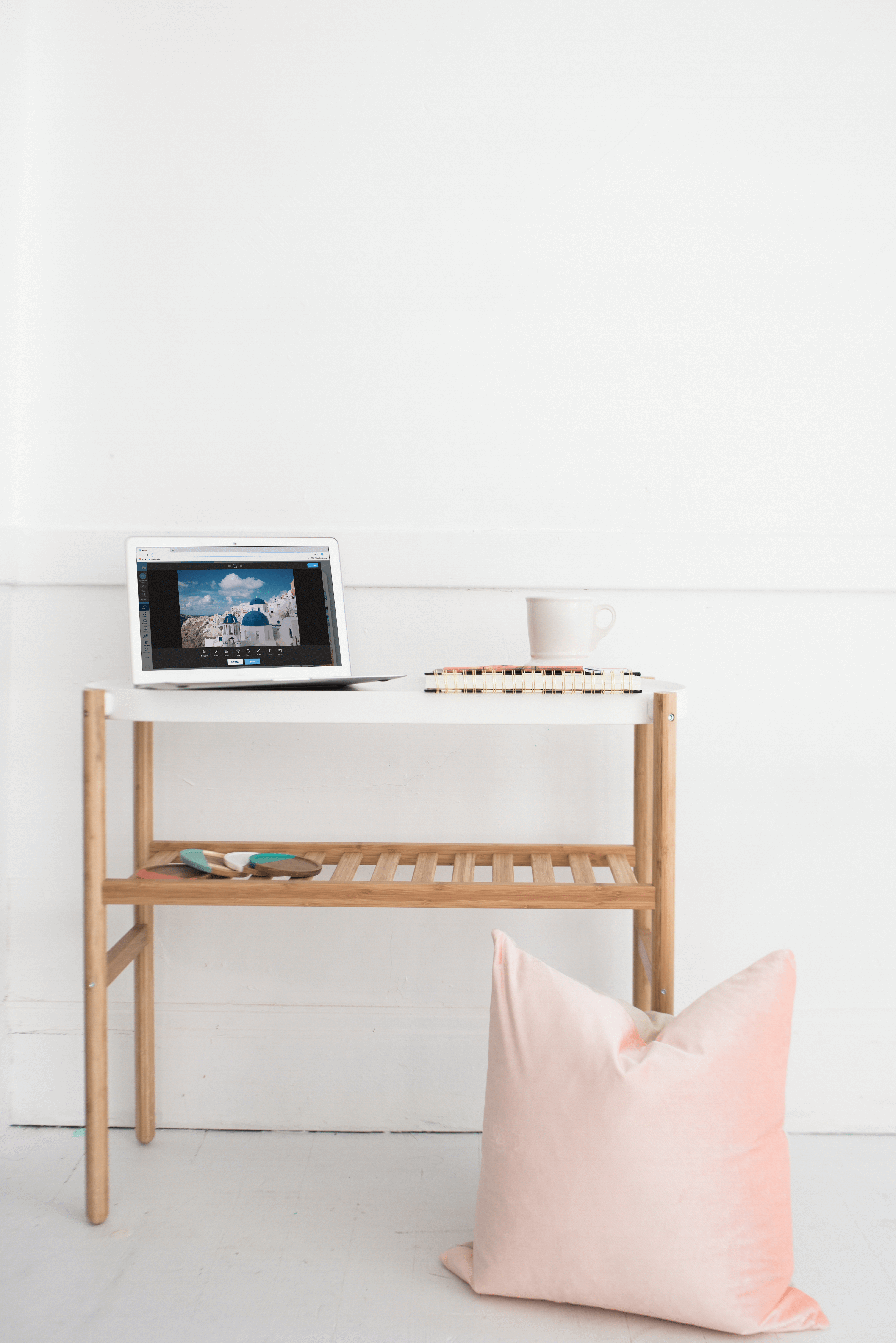 A minimalist desk setup with a laptop, a mug, a notebook, and sunglasses on a wooden table, accompanied by a pink pillow on the floor, ideal for a social media planner | plannthat.com