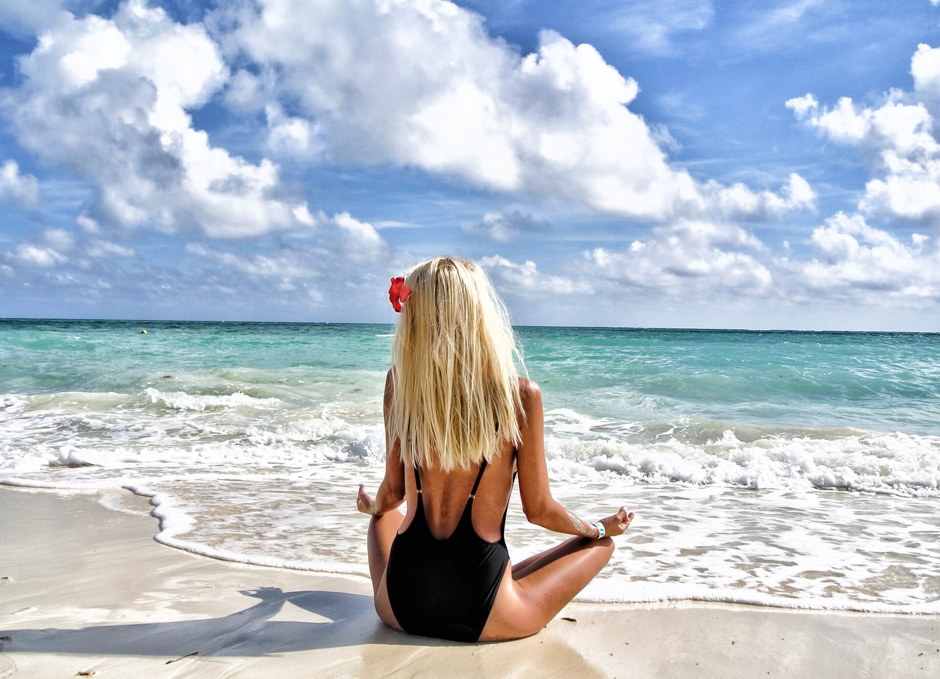 A woman with long blonde hair, wearing a black swimsuit, sits in a meditative pose on a sandy beach facing the ocean, with partly cloudy skies and gentle waves in the background, illustrating a serene moment perfect for a social media planner | plannthat.com