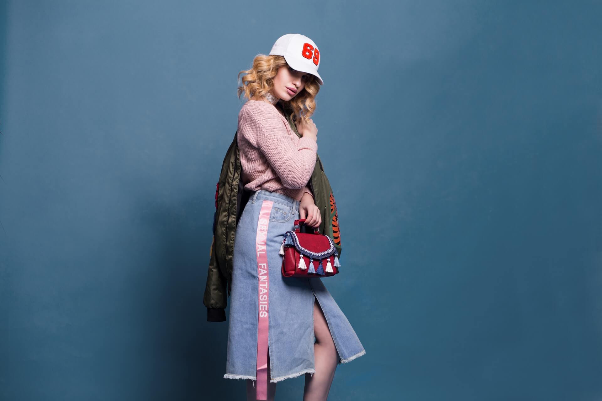 Fashionable woman in a white cap, pink sweater, and blue skirt, holding a red handbag, poses against a blue backdrop | Instagram scheduler | plannthat.com