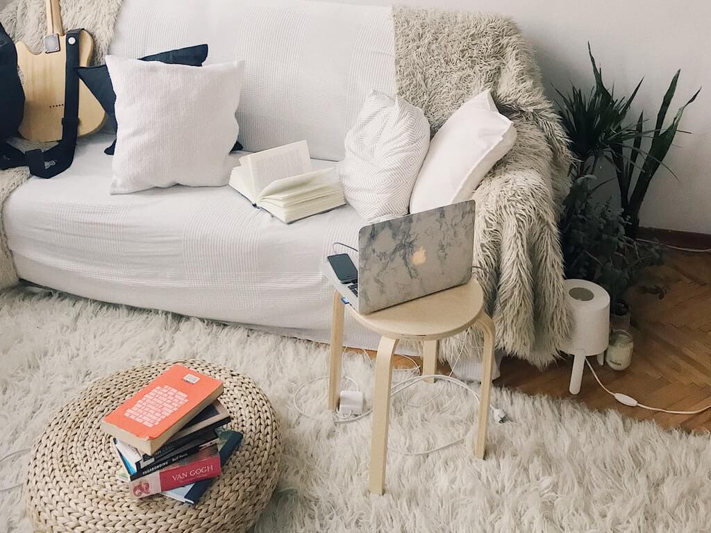 Cozy living room with a white couch, pillows, books, a laptop on a small table, a guitar, and a plant in the corner, creating a comfortable atmosphere perfect for using a social media scheduler | plannthat.com