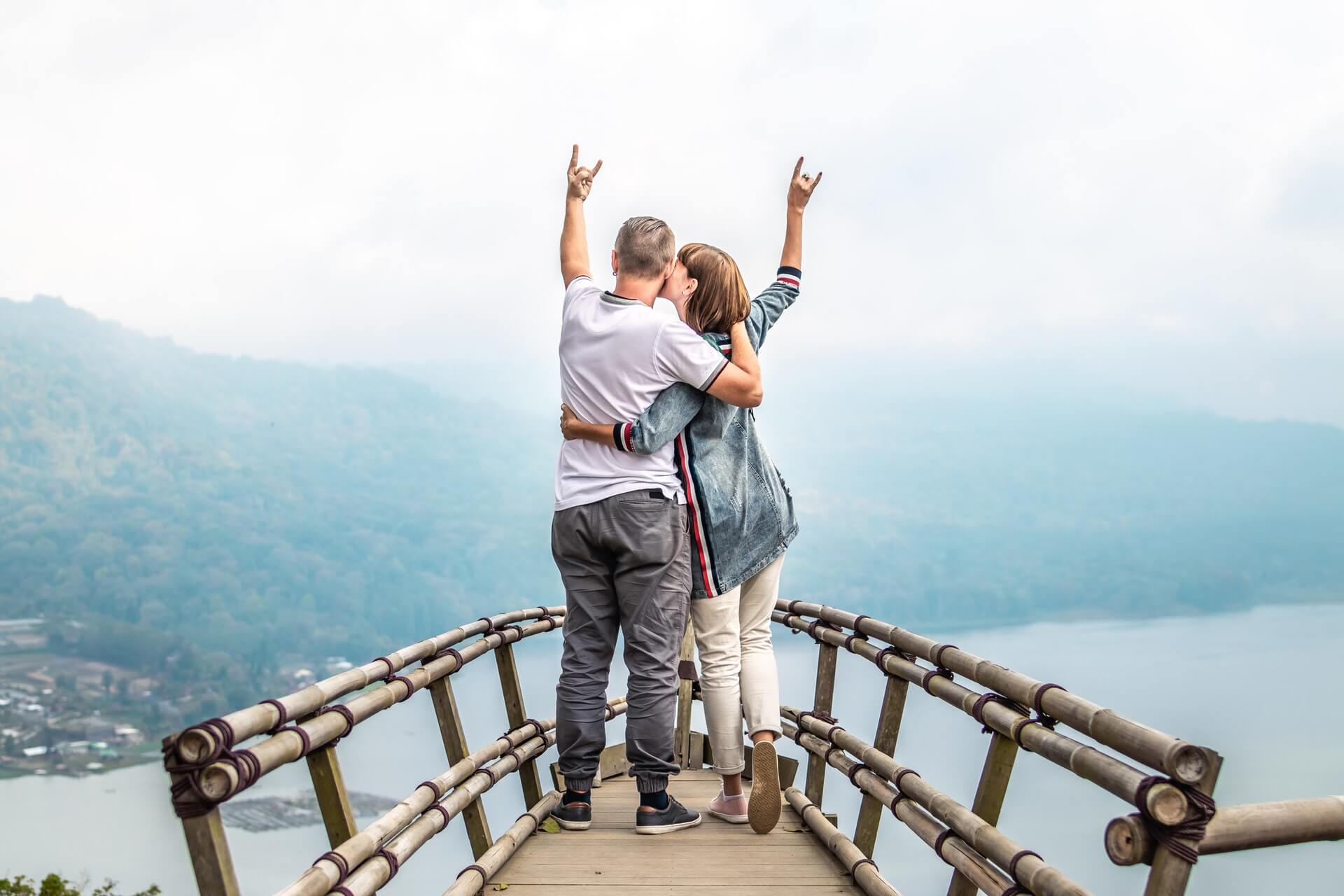 A couple stands on a wooden bridge overlooking a scenic landscape with mountains and a lake, embracing and raising their hands in a celebratory gesture, enjoying the view and the moment together | Instagram scheduler | plannthat.com