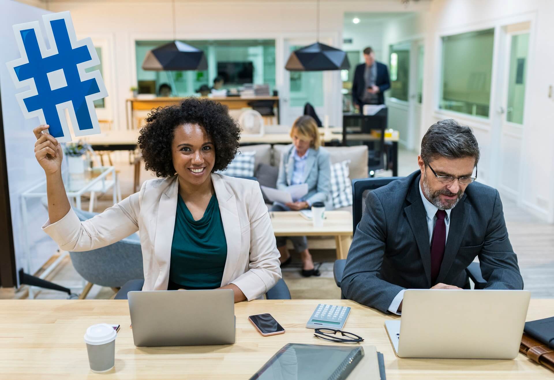 A woman holds a large blue hashtag symbol in an office, smiling at the camera, while a man works on a laptop beside her and other people are in the background, illustrating a collaborative environment for a social media planner | plannthat.com